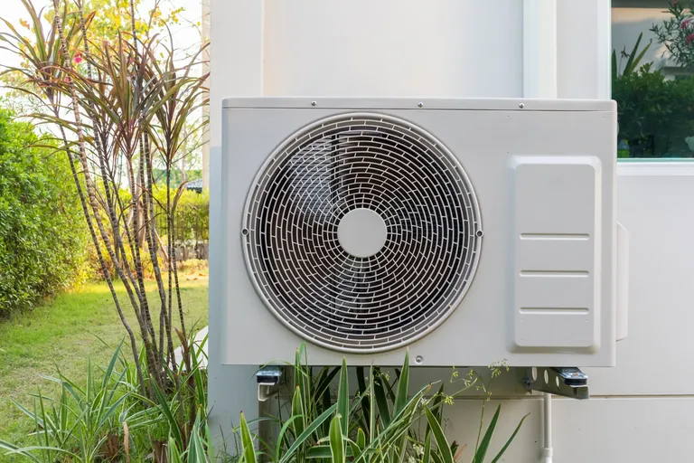Outdoor air conditioning unit mounted against a white wall in a garden