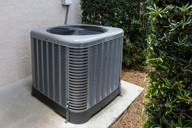 Grey outdoor air conditioning unit installed near bushes